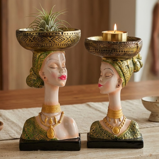 Two decorative female bust candle holders with gold and green patterns, one holding a small plant and the other a lit candle, displayed on a stone table in a warm indoor setting.